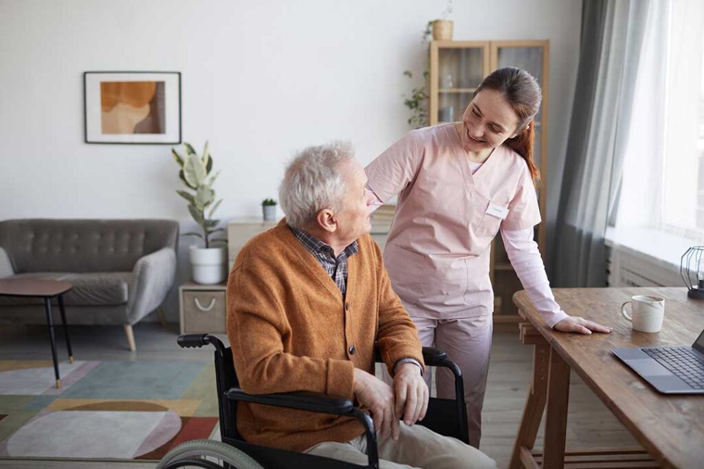 An older adult talking with a caregiver at a table in a bright shared living area.